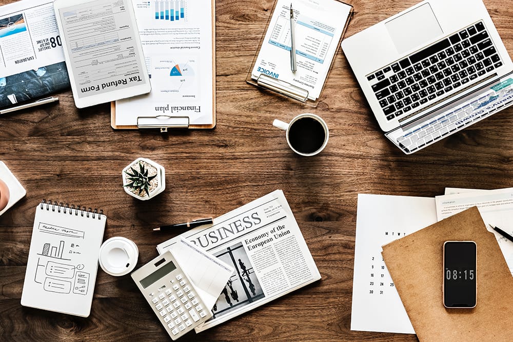 Uma mesa com diversos itens sobre ela. Notebook, jornal, planilhas, gráficos, blocos de notas, calculadora, tablet e uma xícara de café. Representa todos os cuidados em análises de dados na hora de criar um planejamento financeiro
